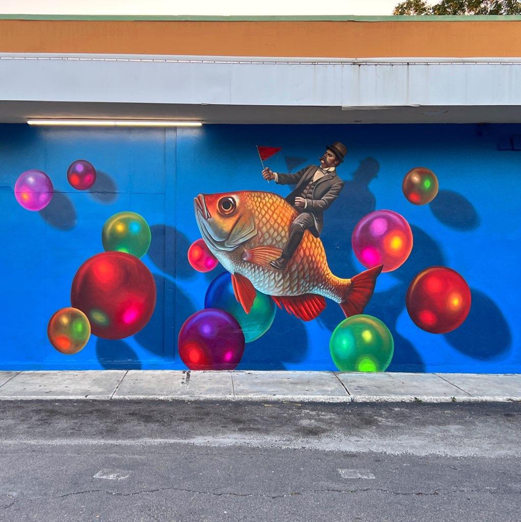 Outdoor mal of a dapper man riding a goldfish among multi colored bubbles.