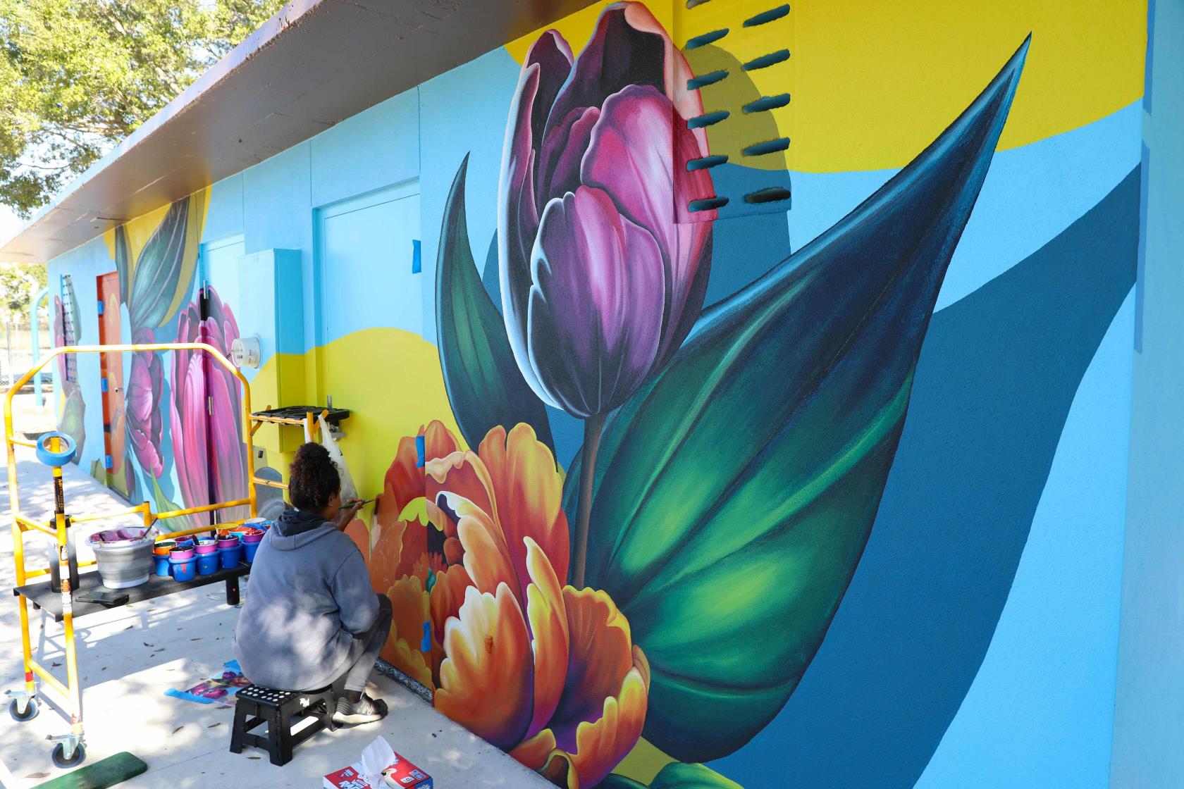 Artist painting a floral mural on the outside of a small building.