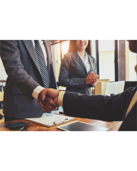 Two men shaking hands over a desk with a woman standing next to them, all in business attire.